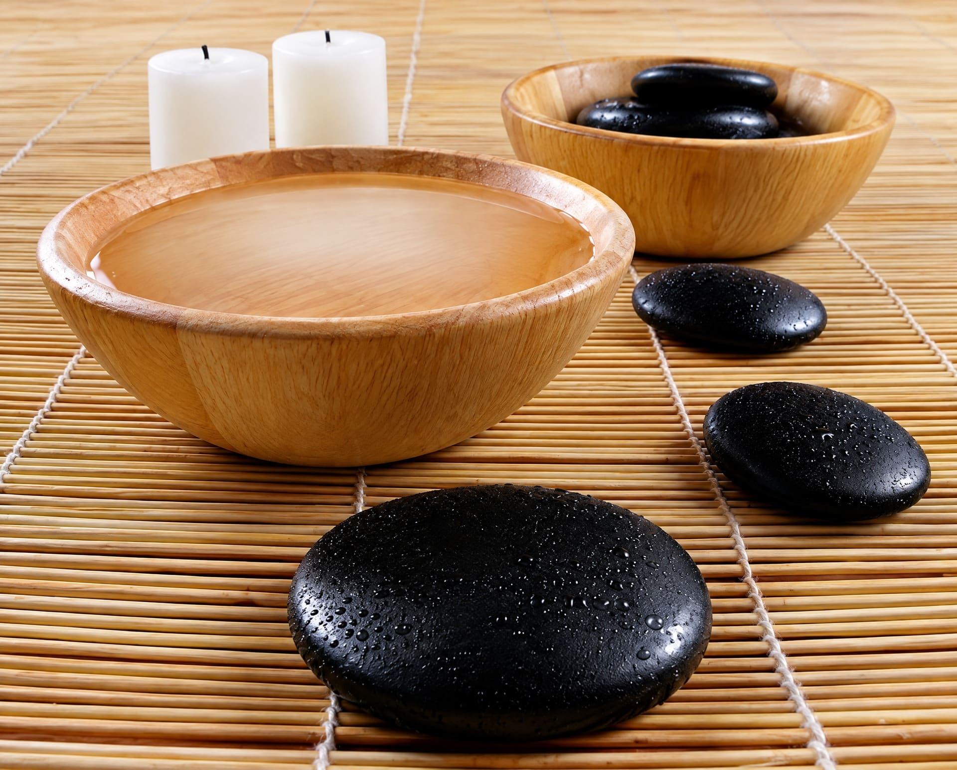 Houten schalen met water en warme massagestenen naast twee kaarsen op een bamboemat